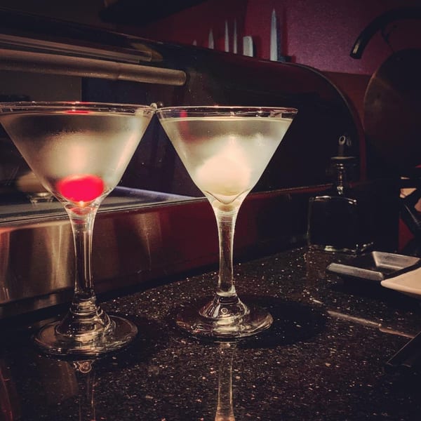 Two martini glasses, one with a red cherry and the other with an olive, on a dark bar.