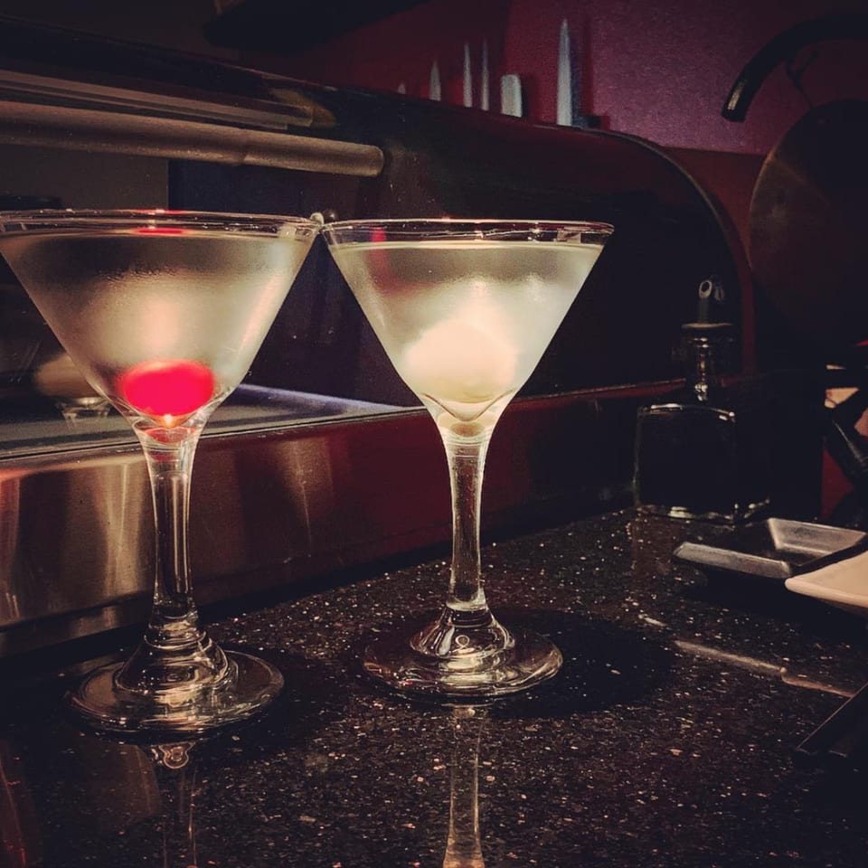 Two martini glasses, one with a red cherry and the other with an olive, on a dark bar.