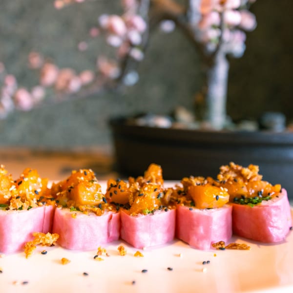 Pink sushi rolls topped with orange mango, black and white sesame seeds, and crispy bits.