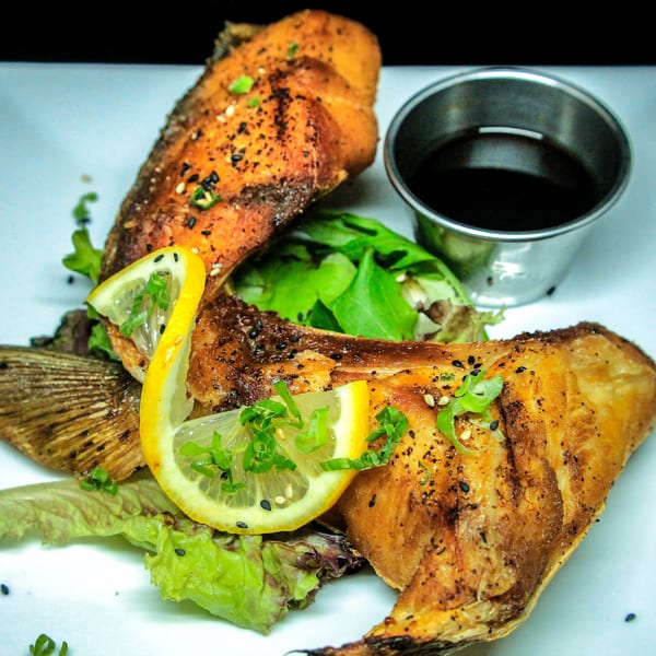 Grilled fish with lemon slices, green onions, sesame seeds, and a side of dark sauce.